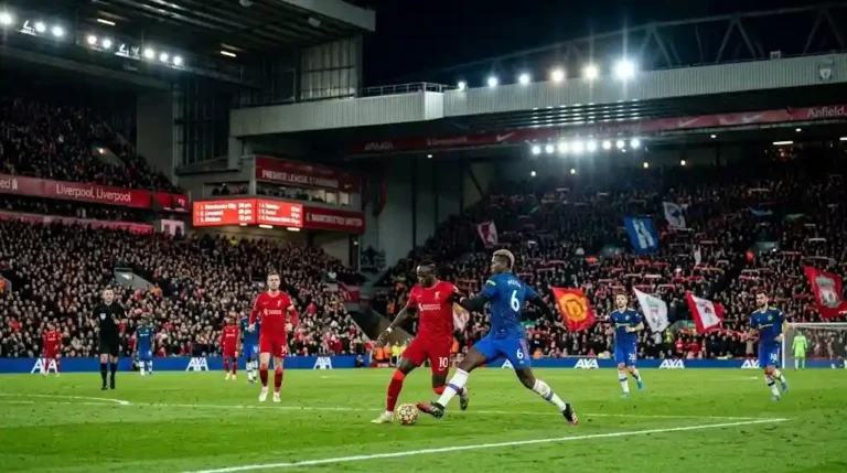 Partido de fútbol con jugadores disputando el balón frente a la pantalla con las posiciones de premier league.